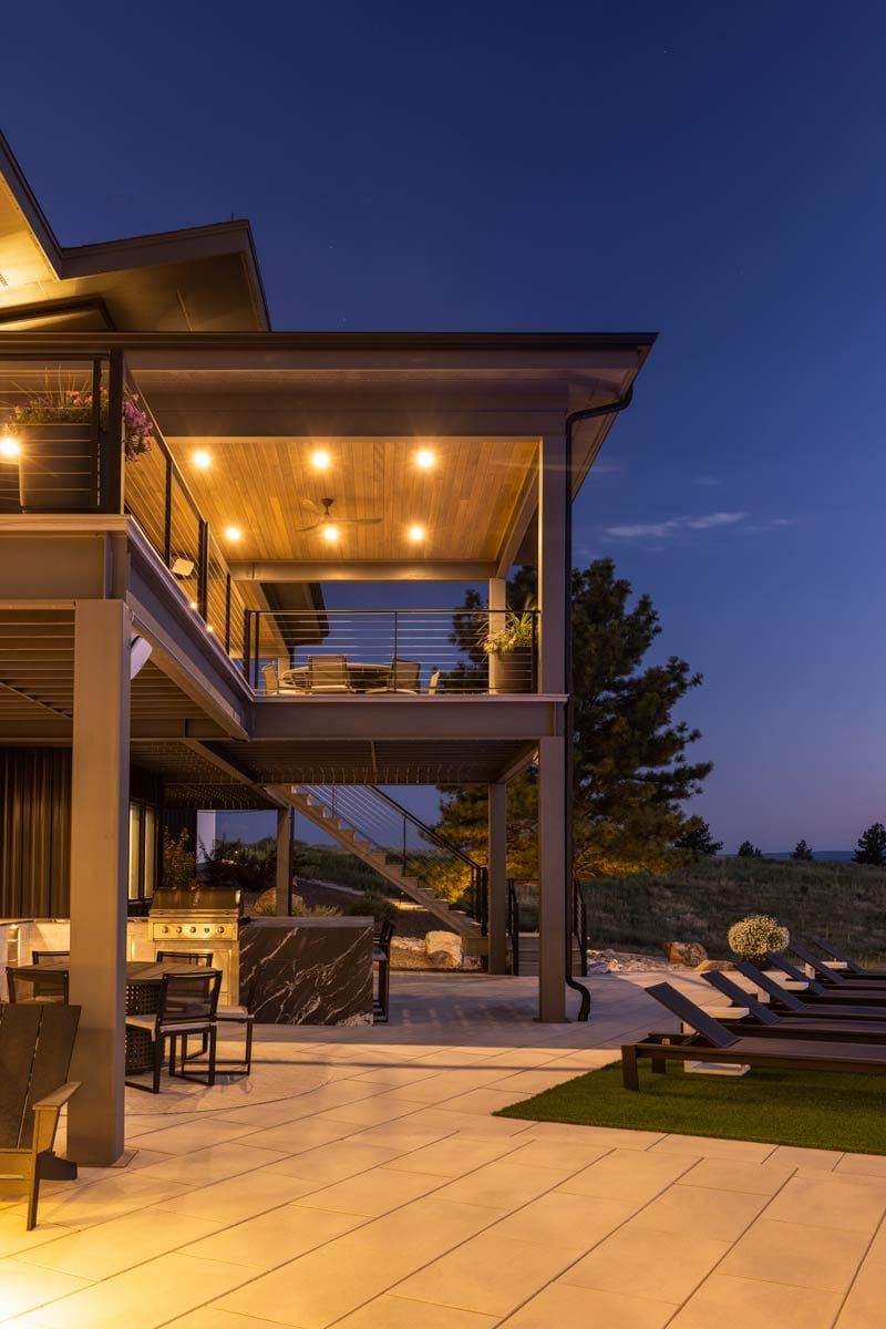 Multi-level modern home exterior at dusk featuring a covered upper deck with lights, an outdoor kitchen, and a lower patio with lounge chairs.