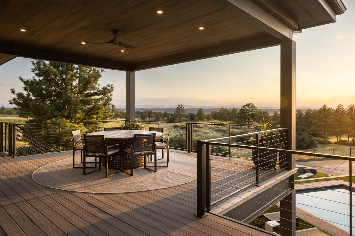 Covered deck with dining set and ceiling fan, overlooking a wooded landscape at sunset.