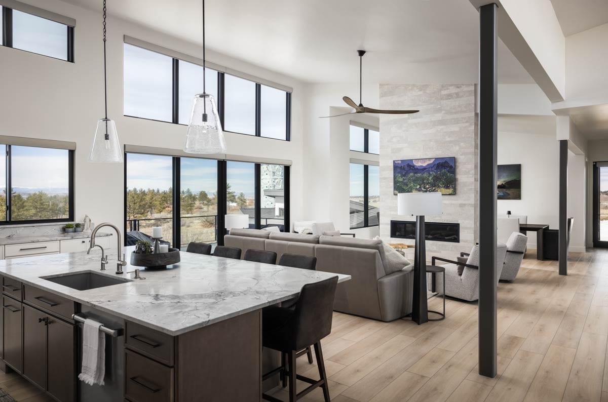 Open concept living area with kitchen island, seating, fireplace, and large windows. Wood floors and ceiling fan visible.